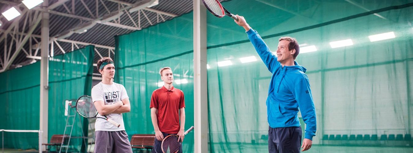 Tennisvalmentaja opettaa kahdelle nuorelle miehelle lyöntitekniikkaa.