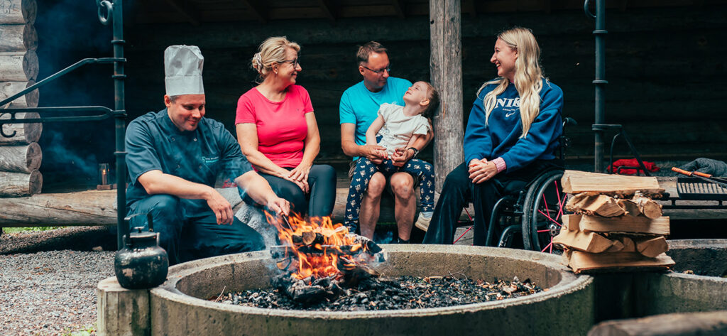 Pajunpesä-laavulla kokki kohentaa nuotion tulta ja kaksi naista, mies ja lapsi istuskelevat hymyileväisinä.