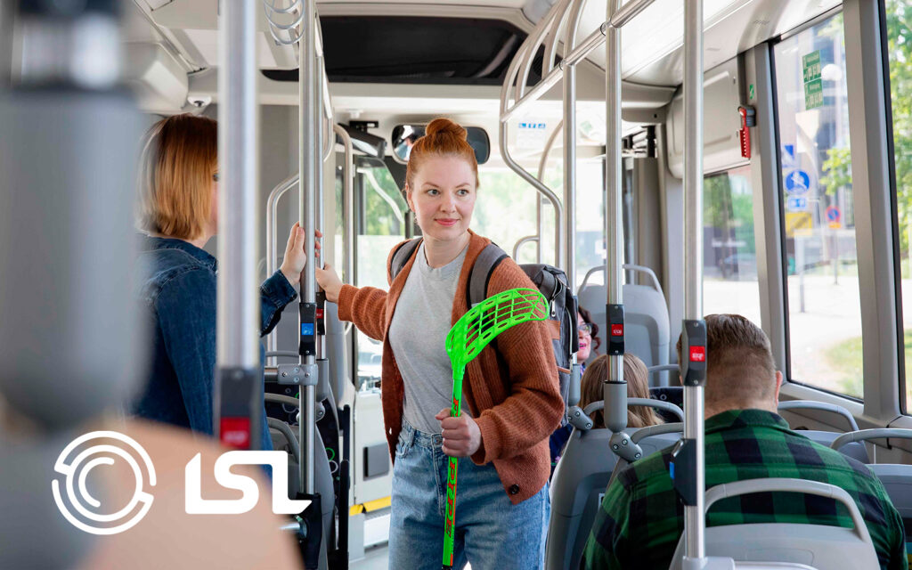 Linja-autossa nainen seisoo käytävällä pidellen kädellä kiinni sählymailasta ja toisella kädellä kaiteesta.