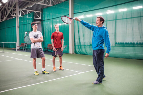 Tennishalli Pajulahdessa. Tennisopettaja ohjeistaa mailan asentoa kahdelle nuorelle miehelle.