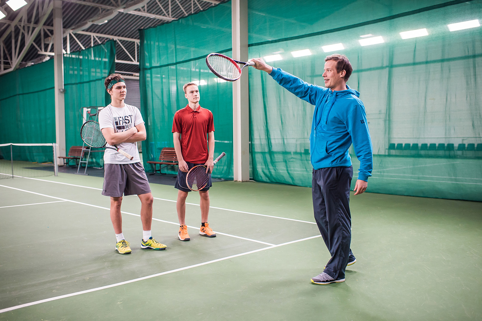 Tennishalli Pajulahdessa. Tennisopettaja ohjeistaa mailan asentoa kahdelle nuorelle miehelle.
